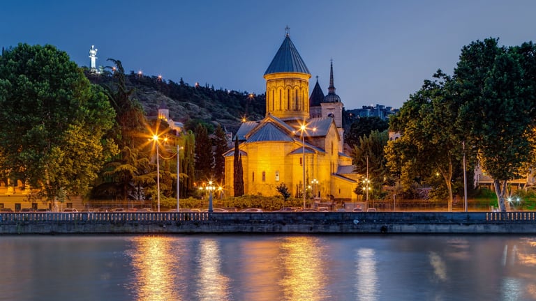 Illuminated Orthodox church with golden dome reflected in river at twilight, surrounded by trees and street lights