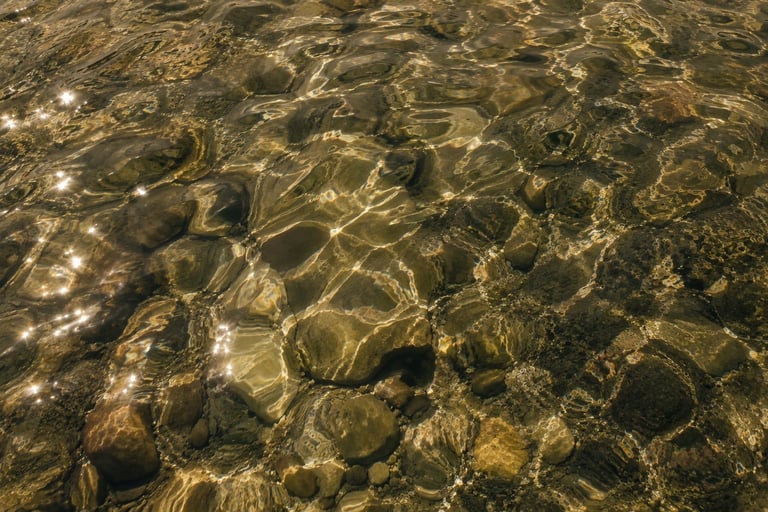 Clear shallow water with sunlight reflecting off rocks and pebbles on the riverbed or beach floor