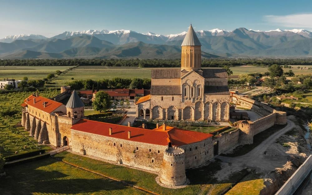 Aerial view of Akdamar Church with golden bell tower, surrounded by fortress walls with red roofs, set in a green valley with snow-capped mountains in background