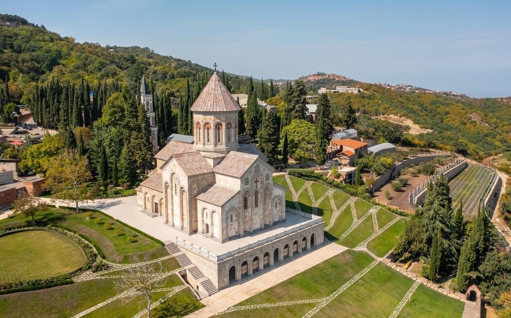 Aerial view of a stone Armenian church with conical dome surrounded by manicured gardens and cypress trees on a hillside