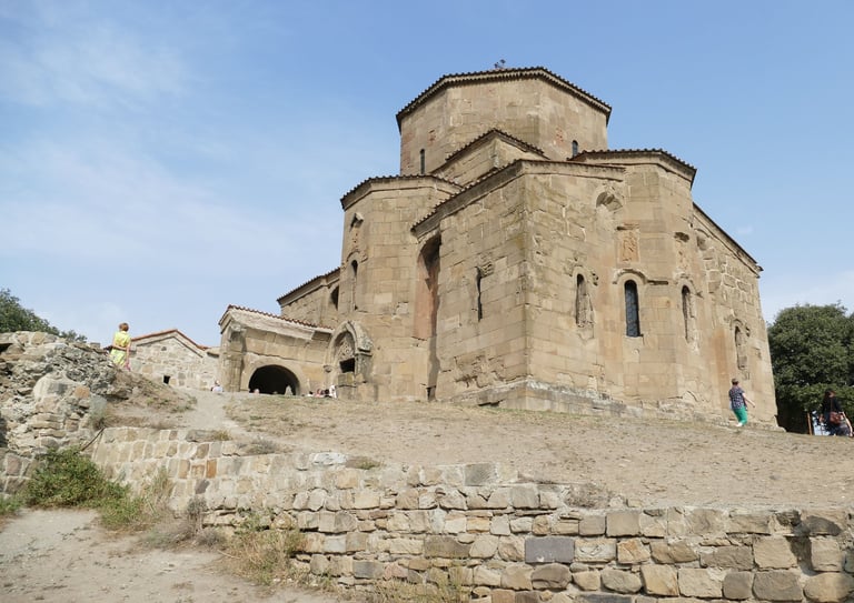 Ancient stone church with two domes on a hillside, surrounded by stone ruins and greenery under blue sky
