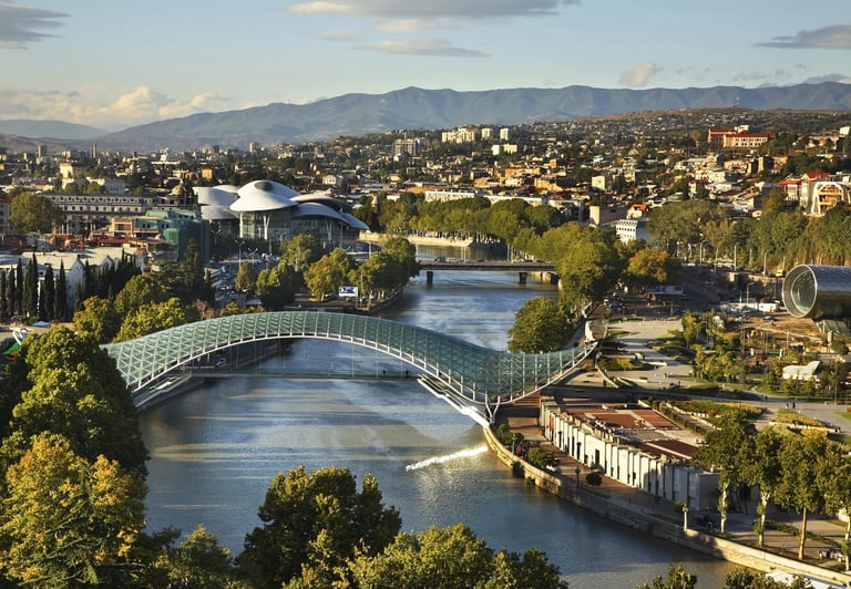 Aerial view of a modern pedestrian bridge spanning a river with city buildings and mountains in the background