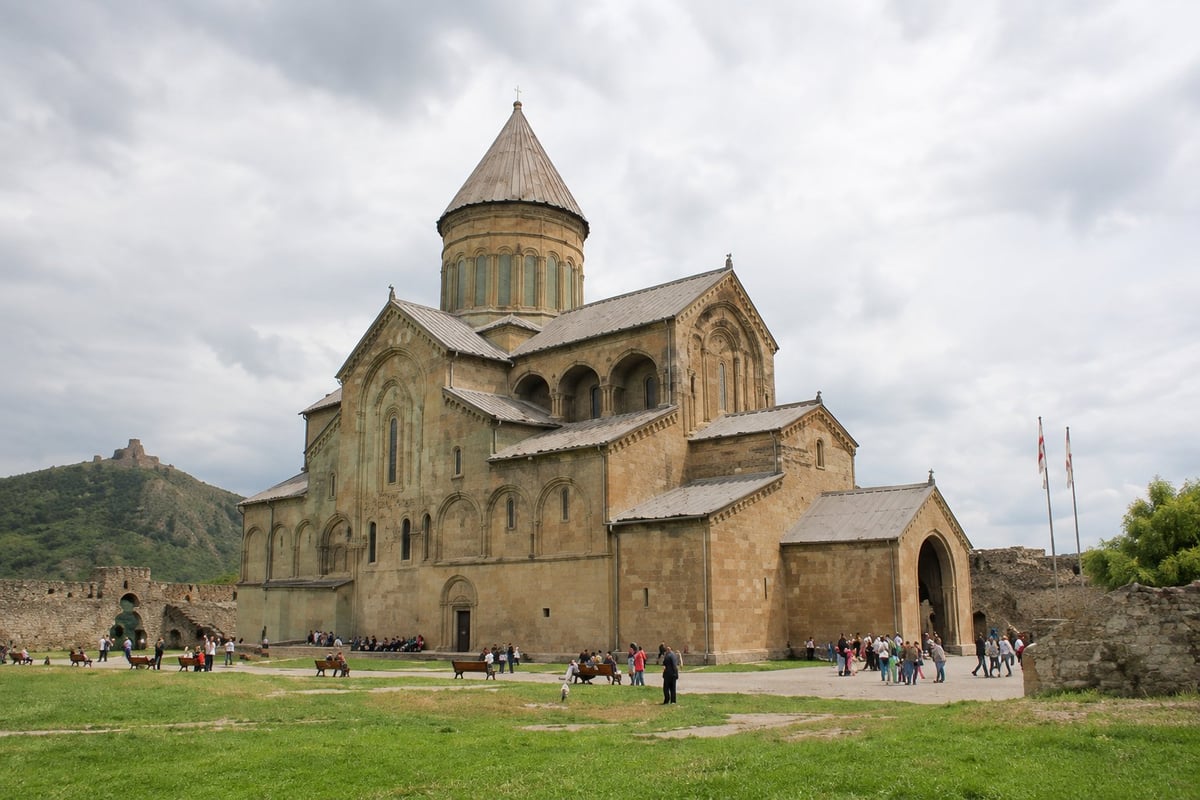Ancient stone church with golden domed roof and conical tower, surrounded by visitors on green grounds under cloudy sky