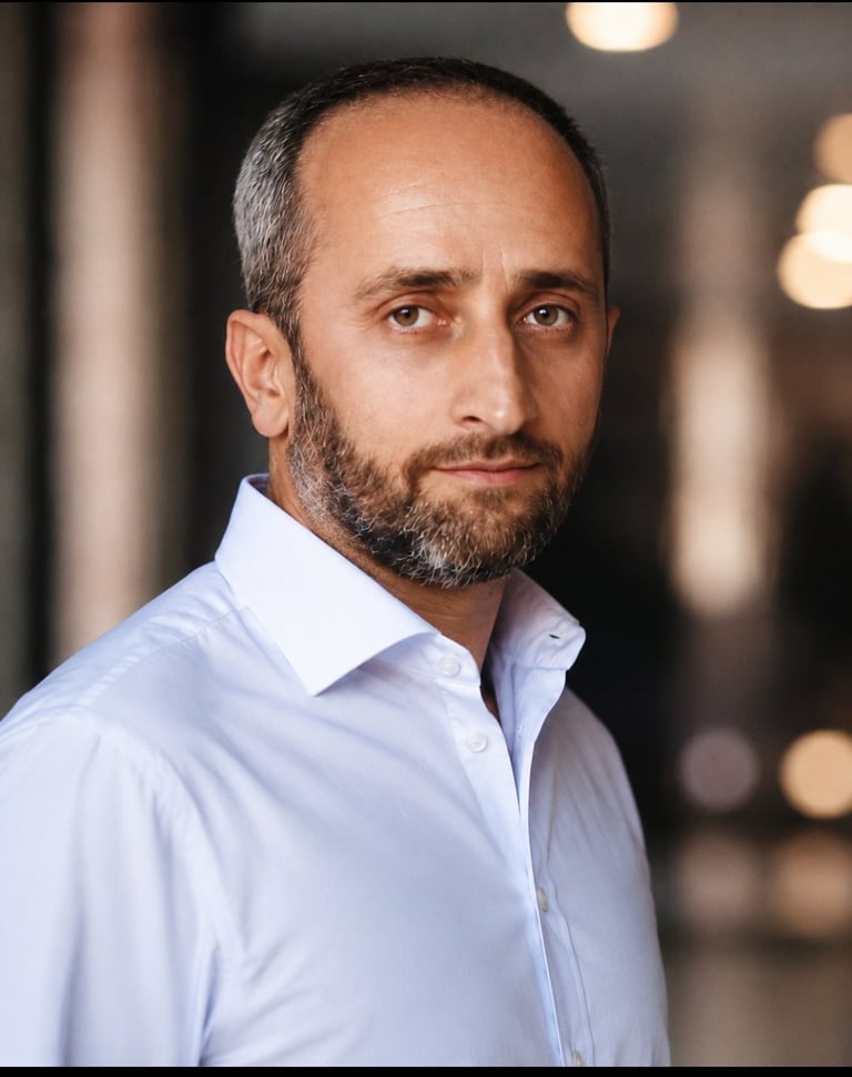 Professional headshot of a bearded man with dark hair wearing a light blue button-up shirt against a blurred indoor background