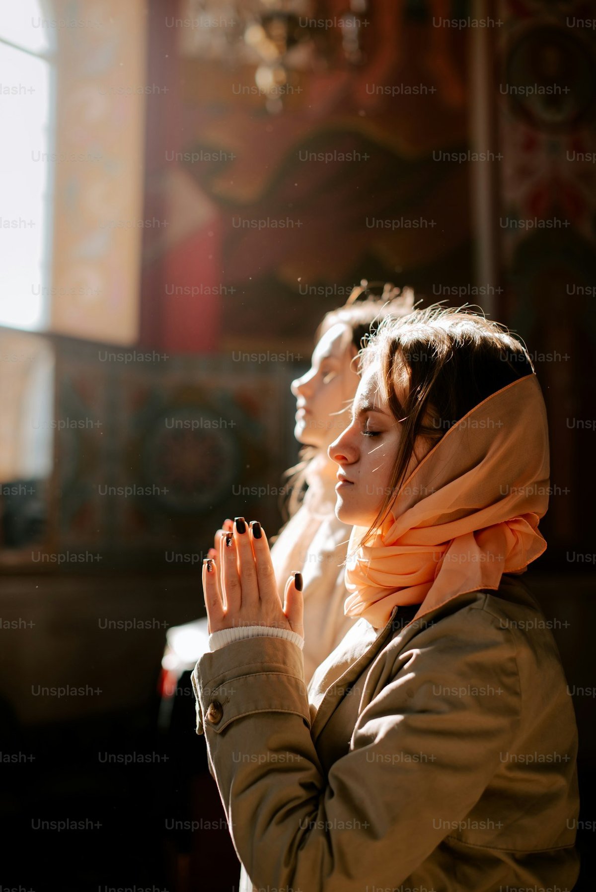 Woman wearing orange headscarf and beige jacket praying indoors with warm sunlight streaming across her face
