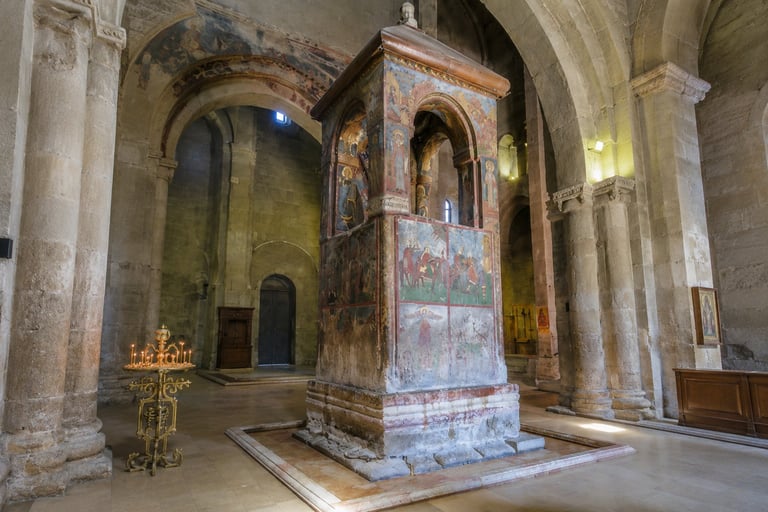 Interior of ancient church with stone columns, ornate central altar structure, faded religious frescoes, and golden candleholder