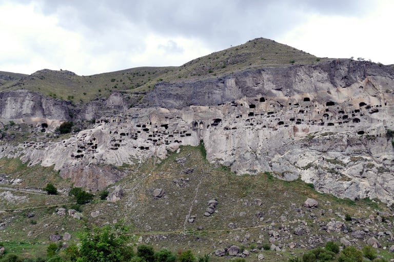 Hillside with ancient cave dwellings carved into light-colored rock faces beneath a green mountain slope under cloudy sky